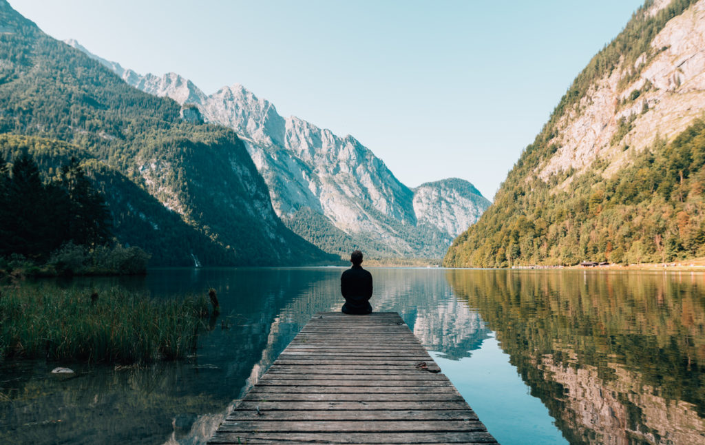 A person sits at the edge of a dock with their back turned to the viewer. The lake is surrounded by mountains.