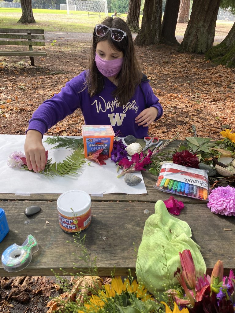 Picture of young girl with brown hair sitting at a picnic table using flowers and plants to create a self portrait